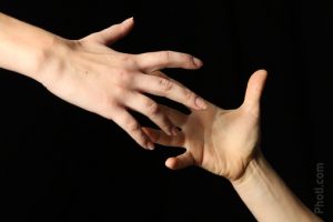 The background is black and there are two hands -- light skinned -- one from the left and one from the right whose fingers are reaching for each other.