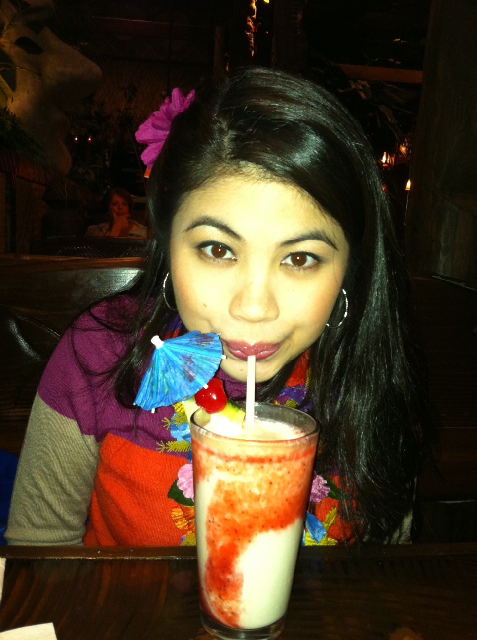 Woman with long dark is drinking a colorful drink that has a blue paper umbrella in it.