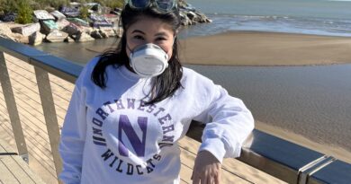 Picture of a young woman with dark hair and a white P100 mask smiling at the camera. She carries a cane. The background is a lake and sand on a sunny summer day.