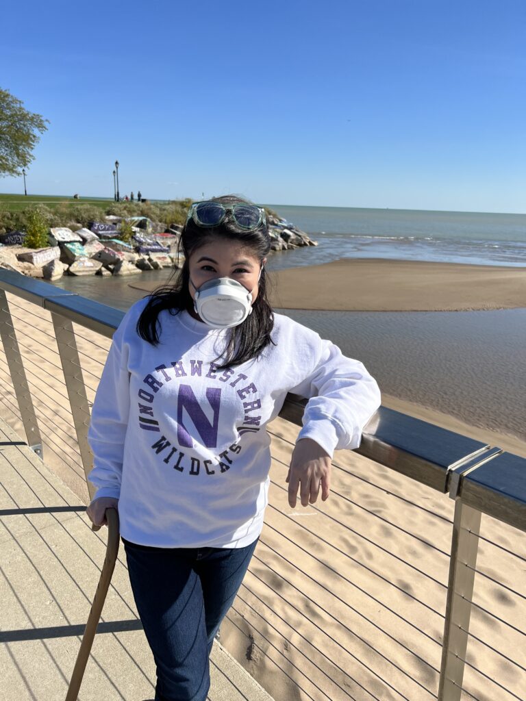 Picture of a young woman with dark hair and a white P100 mask smiling at the camera. She carries a cane. The background is a lake and sand on a sunny summer day.