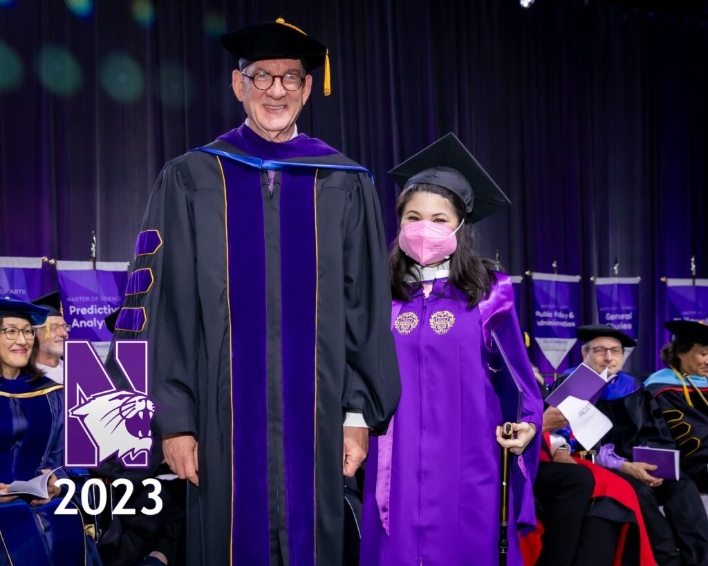 Image shows a 6-something White man with a black graduation cap and a black gown with purple regalia. Standing next to him is a 20-something looking Asian - Hispanic-looking woman. She is wearing a black graduation cap, hot pink N95 mask, and a purple graduation gown that has gold regalia on it. She is also carrying a black cane. They are both smiling at the camera.