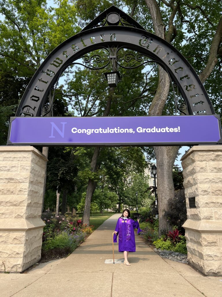 Picture of two Asian-looking women--daughter and mom in front of a purple rock. The painted rock says, "Congrats, graduates!" and has a wildcat footprint. Daughter is carrying a brown cane and wearing a purple graduation gown. She has long, black hair. Mother has grey hair and is wearing all white.