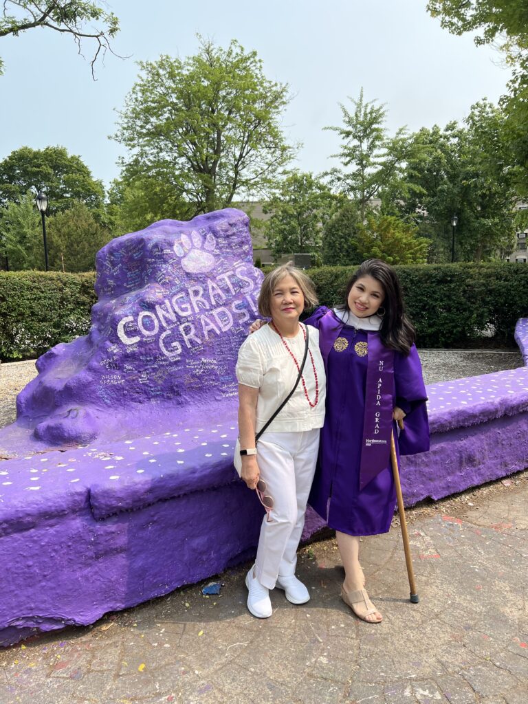 Picture of two Asian-looking women--daughter and mom in front of a purple rock. The painted rock says, "Congrats, graduates!" and has a wildcat footprint. Daughter is carrying a brown cane and wearing a purple graduation gown. She has long, black hair. Mother has grey hair and is wearing all white.