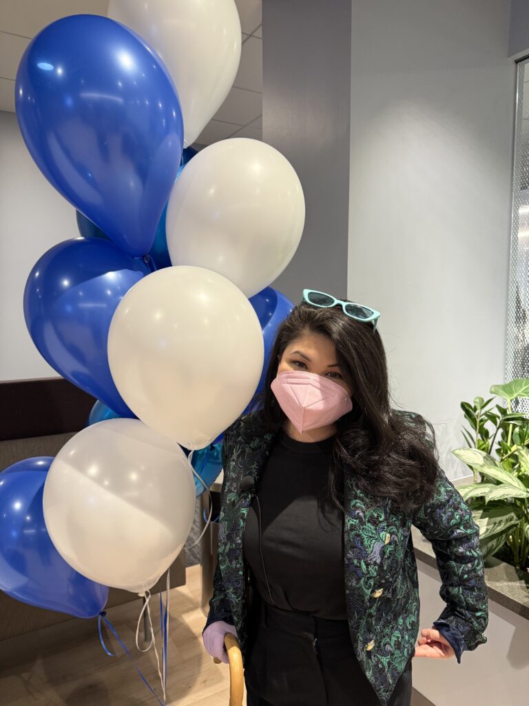 Picture of Asian/Latina looking woman with long black hair, blue sunglasses on top of her head, green and blue brocade blazer, black dress pants wearing a pink face mask smiling. She is standing next to blue and white balloons.