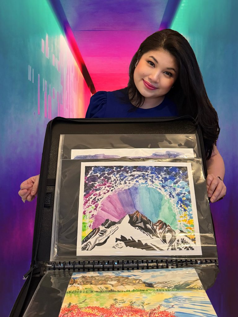 Picture of Asian / Hispanic looking woman with dark hair smiling at the camera while holding art portfolio of her watercolor paintings. She is wearing a bright violet dress. The painting facing forward is of a white mountain range against a colorful sky featuring the Milky Way arc, Northern Lights and an airglow. There are stars all over the sky. Painting beneath it features another mountain range but on land--not from an aerial view. Mountains are brown with blue sky behind it. A blue lake, green trees and red flowers.